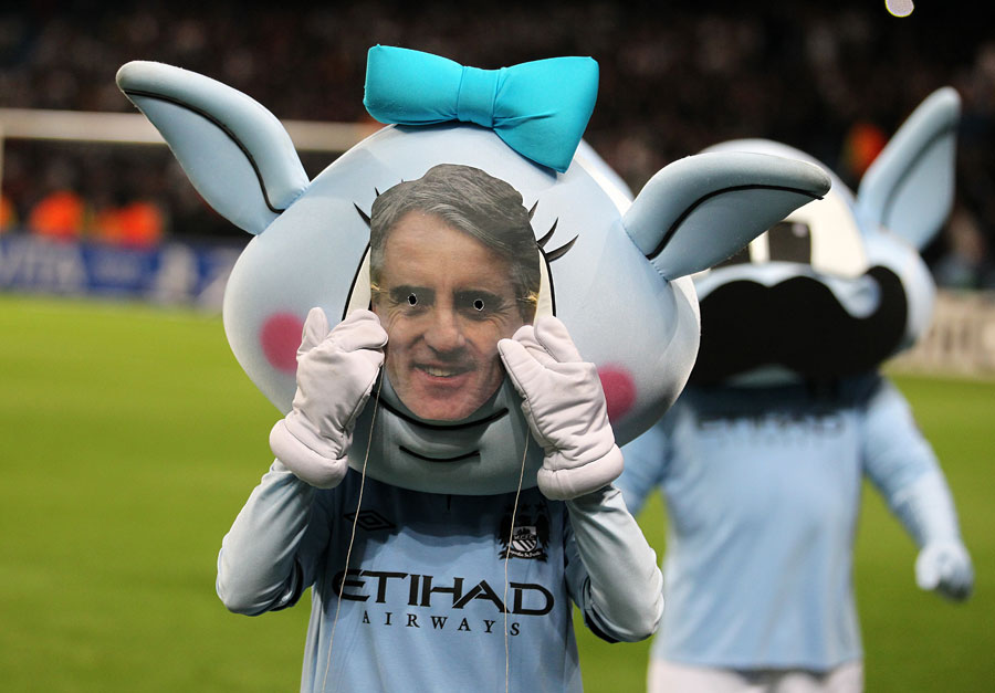 A Manchester City mascot wears a mask of Roberto Mancini